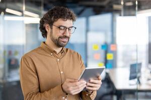 Mature Hispanic man in a smart casual outfit smiles as he focuses on his tablet in a modern office setting. photo