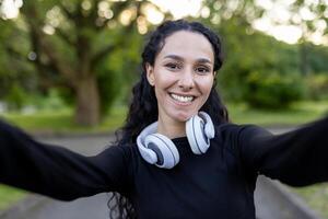 A joyful Hispanic woman wearing headphones takes a selfie outdoors. She's smiling in a park with a green background, exuding happiness and vitality. photo