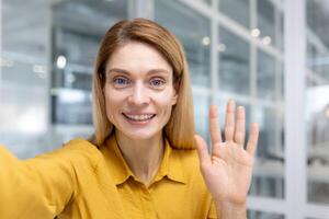 en línea llamar, mujer de negocios dentro oficina a lugar de trabajo mirando a teléfono inteligente cámara utilizando llamada aplicación hablando con amigos y sonriente alegremente. foto