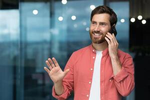 Successful businessman in a red shirt talking on the phone inside the office, a man with a beard at the workplace is talking to a client, a satisfied boss in casual clothes. photo