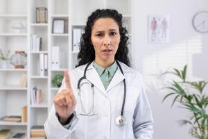 llamada en línea consulta de un doctor, un mujer en un médico blanco Saco mira a el cámara, disgustado y alarmado, menea su dedo, prohíbe el paciente a comer mala calidad comida foto