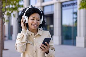 joven hermosa africano americano mujer caminando en el ciudad vistiendo auriculares sonriente escuchando a música y audio libros, mujer utilizando teléfono inteligente solicitud para en línea podcasts foto