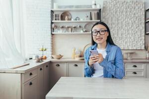 un contento joven asiático Adolescente niña es sentado a hogar en el cocina, participación un teléfono en su manos y enviando un mensaje a su amado uno, un novio. mirando a el cámara, sonriente. foto