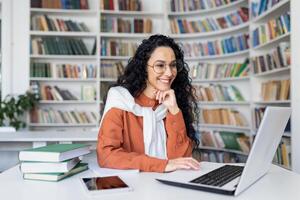 hembra estudiante con ordenador portátil leyendo en línea curso, latín americano mujer sonriente y satisfecho con independiente en línea aprendizaje sentado dentro Universidad instalaciones en biblioteca. foto
