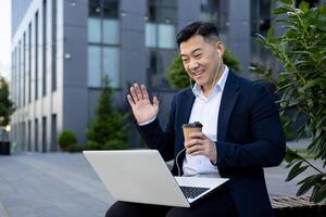 sonriente joven asiático hombre en un negocio traje sentado en un banco cerca un oficina edificio vistiendo auriculares, participación un taza de café, hablando en un llamada en un computadora portátil, diciendo Hola a el cámara. foto