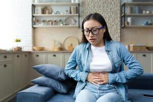 Stomach disorder. A young beautiful Asian woman holds her stomach with her hands, feels severe pain. Sitting at home on the sofa. photo