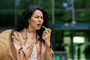 Upset female holding small green bottle and spraying throat spray into mouth to alleviating pain outdoors. Lady wearing light brown coat and shirt with brown spots on background of glassy building. photo