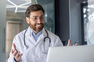 de cerca foto de un joven sonriente especialista médico vestido en un túnica sentado en un hospital oficina en frente de un ordenador portátil y hablando en línea vía llamar, consultante un paciente remotamente