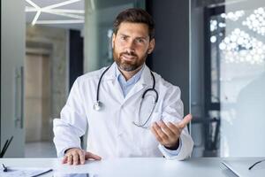 Close-up portrait of a young specialist doctor who talks online via link, sits in the hospital at the table in front of the camera and consults remotely. He gestures with his hands and explains. photo