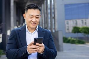 confidente asiático empresario en un traje comprometido con su teléfono inteligente en un ciudad calle, radiante positividad foto