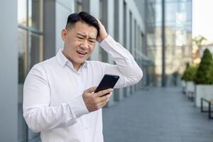 trastornado joven asiático hombre en pie cerca un oficina edificio, mirando frustrado a el teléfono pantalla y participación su cabeza en inquietud. foto