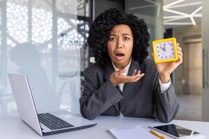 Overworked woman working late, businesswoman looking at camera upset showing clock, female worker with laptop in middle of office tired. photo