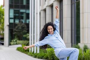 An exuberant woman dances freely outside a modern building, embodying joy and liberation. She celebrates life with her dynamic movement, invoking feelings of happiness and spontaneity. photo