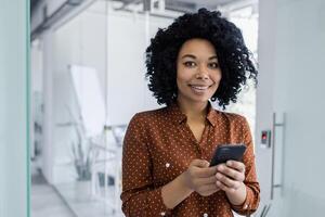 confidente africano americano mujer de negocios utilizando su teléfono inteligente con un agradable sonrisa, en pie en un brillante moderno oficina. profesional atuendo y positivo lugar de trabajo energía. foto