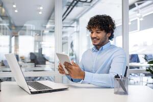 un profesional hombre multitarea con un tableta y ordenador portátil en un brillante, contemporáneo oficina espacio, exudando eficiencia y productividad. foto