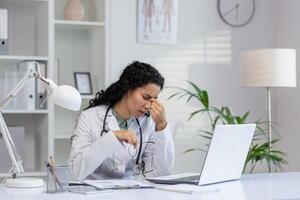 Exhausted Latina doctor in white coat rubbing eyes at desk with laptop, showing signs of stress and burnout in medical office. photo