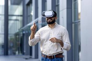 A young man with a beard is standing outside a modern building, wearing a virtual reality headset and gesturing with his hands as if interacting with the virtual environment. The image evokes a sense of immersion and technological advancement. photo