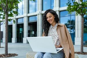 Smiling Hispanic woman sitting outdoors with a laptop, working happily near modern office building. She exudes professionalism and confidence while enjoying a sunny day. photo