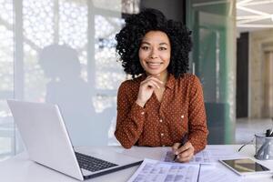 Latino woman in polka dot blouse filling out tables by hand while working at technically equipped desk. Motivated office administrator placing order to providing company with necessary supplies. photo