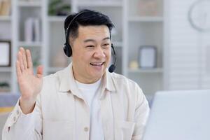 Cheerful Asian man using a headset, engaging in an online conversation from his home office. His expression is friendly and welcoming, suggesting a professional yet casual remote work environment. photo