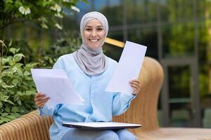 A professional Muslim woman with a headscarf holding papers, exhibiting confidence and a warm smile, sitting on a bench outdoors. photo