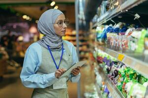 mujer vendedor en súper mercado en hijab con tableta comprobación productos utilizando bolsillo computadora, musulmán mujer cerca estantería con productos y bienes foto