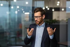 Friendly and smiling businessman in office recording audio message, man using smartphone app for communication, investor in business suit near window inside building. photo