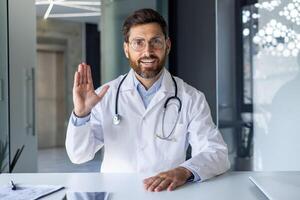 Portrait of a young male doctor in a white coat and with a stethoscope sitting at a table in a hospital, chatting online, greeting and waving at the camera. photo