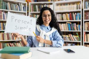Cheerful educator teaching the present simple tense, using a whiteboard in front of a bookshelf-filled library. photo