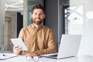 Portrait of a young male programmer working in the office at a laptop and holding a tablet in his hands, smiling and confidently looking at the camera. photo