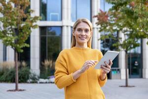 un profesional mujer de negocios en un amarillo suéter usos un digital tableta fuera de un contemporáneo oficina edificio. ella exhibiciones confianza y satisfacción en un corporativo ajuste. foto