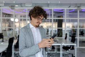 A young man works in an office, stands inside and uses a mobile phone with a smile, typing messages, texting. photo