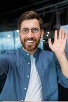 Mature man in office wearing shirt and glasses smiling and looking at smartphone camera, businessman taking selfie and talking on call using smartphone, programmer waving hand greeting gesture. photo