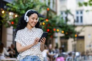 joven hermosa Hispano mujer camina noche ciudad en auriculares, estudiante escucha a música en línea, usos un solicitud en un teléfono inteligente, sostiene el teléfono en su manos, lee mensajes, sonrisas foto