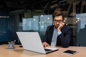 sad and depressed businessman in depression working inside modern office building, man in business suit with glasses and beard using laptop for work, boss disappointed with work result. photo