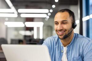 Close-up photo of a smiling young Hispanic man sitting in the office in front of a laptop monitor and working in a headset.