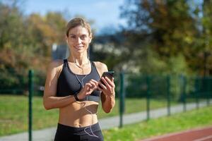 ajuste hembra corredor sonriente mientras ajuste arriba un aptitud aplicación en su teléfono antes de comenzando su rutina de ejercicio en un soleado pista. foto