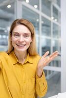 Vertical shot, female employee looking at phone camera waving and smiling, businesswoman talking remotely using smartphone app inside office at workplac. photo