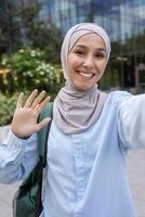 A joyful, professional Muslim woman wearing a hijab takes a selfie with a friendly wave in a modern city environment. photo