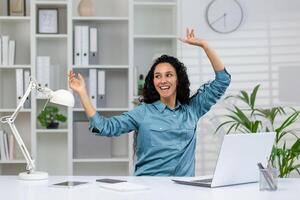 un contento mujer en un azul camisa expresa alegría mientras bailando a su oficina escritorio, rodeado por plantas y un ordenador portátil. foto