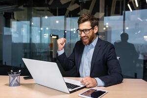 Successful businessman celebrating victory and good business achievement, boss using laptop reading notification from screen and holding hand up smiling triumph gesture. photo