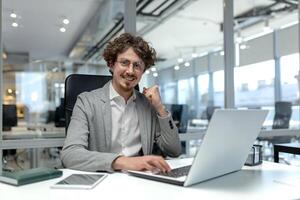 Smiling young businessman with curly hair focused on work at his laptop in a well-lit modern corporate office setting. photo