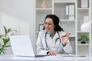 un alegre Hispano hembra médico es visto hablando a un paciente durante un llamada consulta desde su oficina. foto