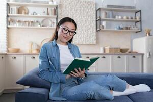 joven hermosa asiático mujer estudiando a hogar, contento hembra estudiante sonriente leyendo libro foto