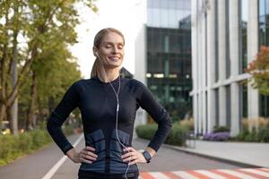 A happy, fit woman with earphones pauses during her urban jog. She's resting with hands on hips as she savors a moment of fitness in a city environment, exuding health and positivity. photo