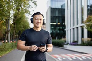 sonriente joven asiático hombre en pie en ciudad calle, corriendo vistiendo auriculares y participación teléfono, mirando lejos. foto