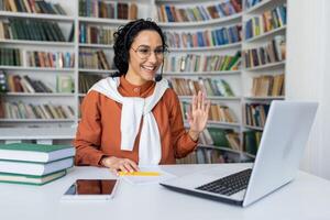 Joyful curly Hispanic girl sitting at a table in front of a laptop in a headset looking at the camera and waving at the camera, office worker in a call center, working call. photo