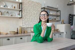 un joven mujer bebidas café a hogar. tiene un descanso. él sostiene un taza en su manos, mira fuera el ventana foto