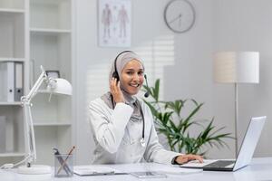 Professional female Muslim physician in hijab engaged in a call consultation, providing medical advice from her office. photo