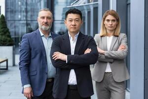 Successful and serious diverse team of three business people, man and woman focused looking at camera with arms crossed, portrait of co-workers outside office building photo